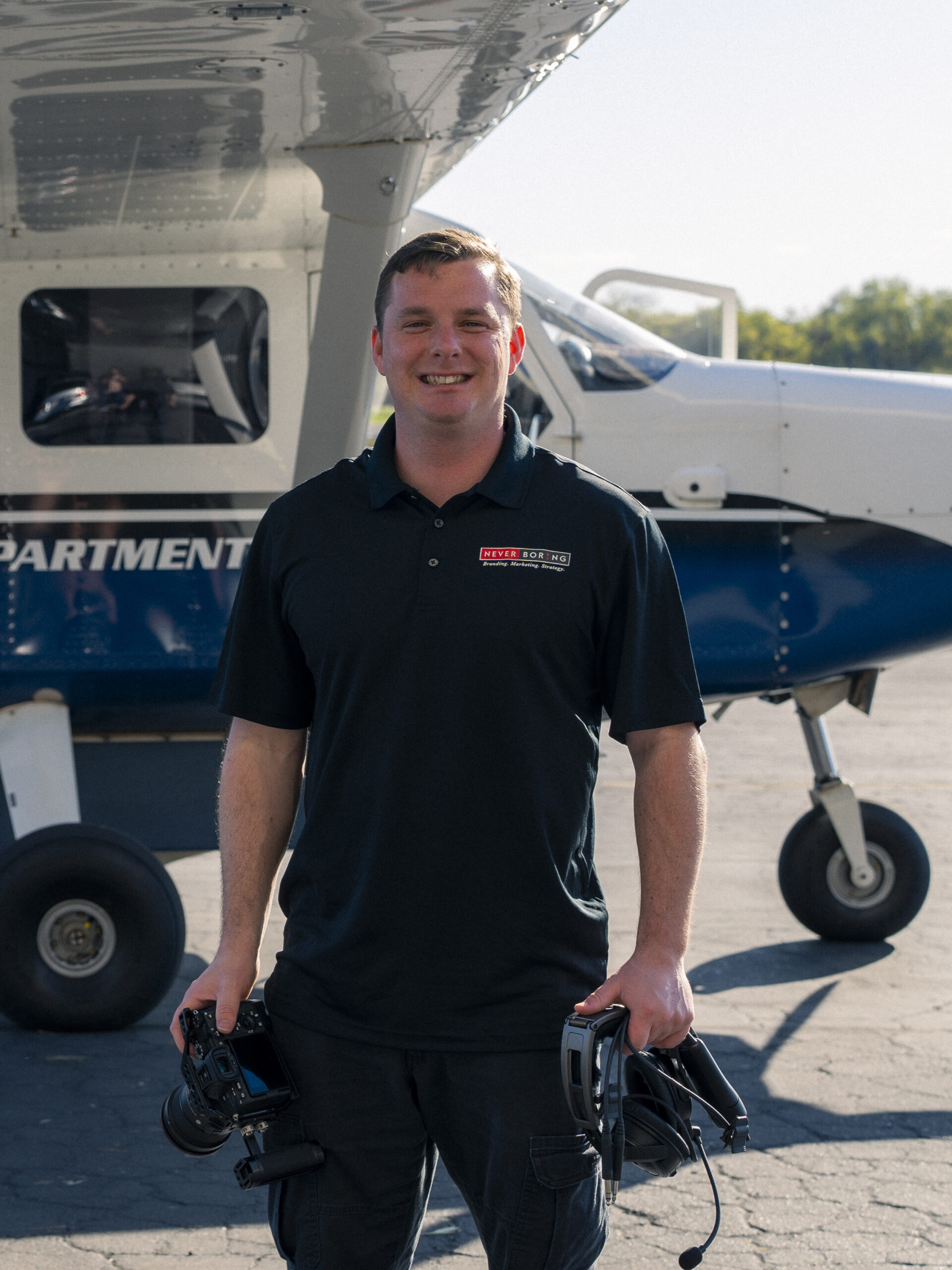 Aaron Davis, Film Specialist standing in front of a small plane holding camera equipment